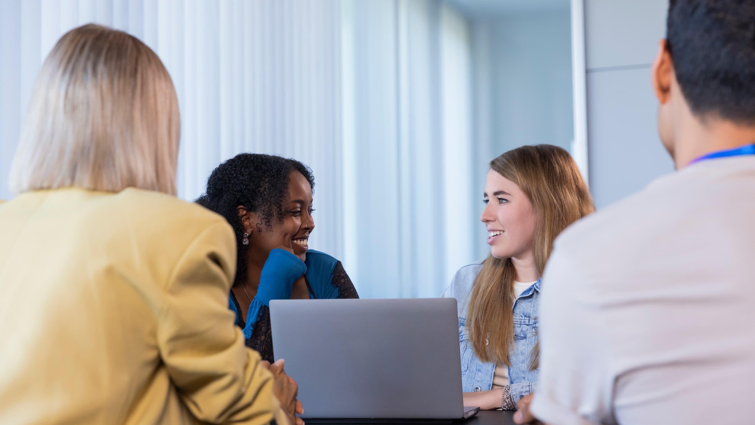 Group of four people in a friendly discussion around a table, with two women in focus smiling across a laptop in a well-lit meeting room.