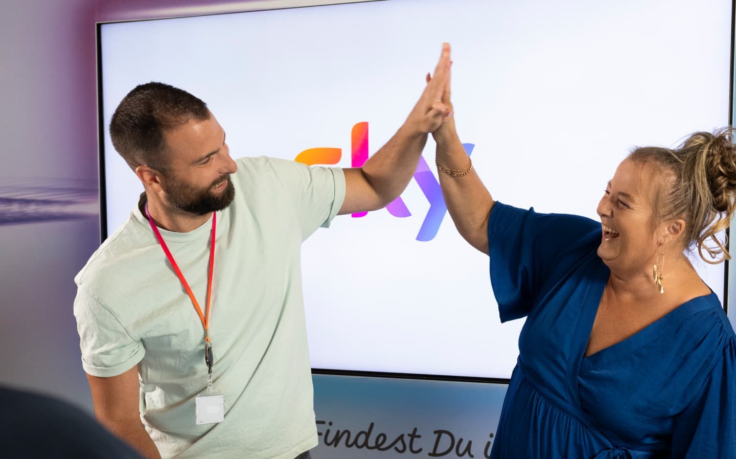 Two colleagues enthusiastically high-fiving in front of a screen displaying the Sky logo, both smiling and wearing lanyards.