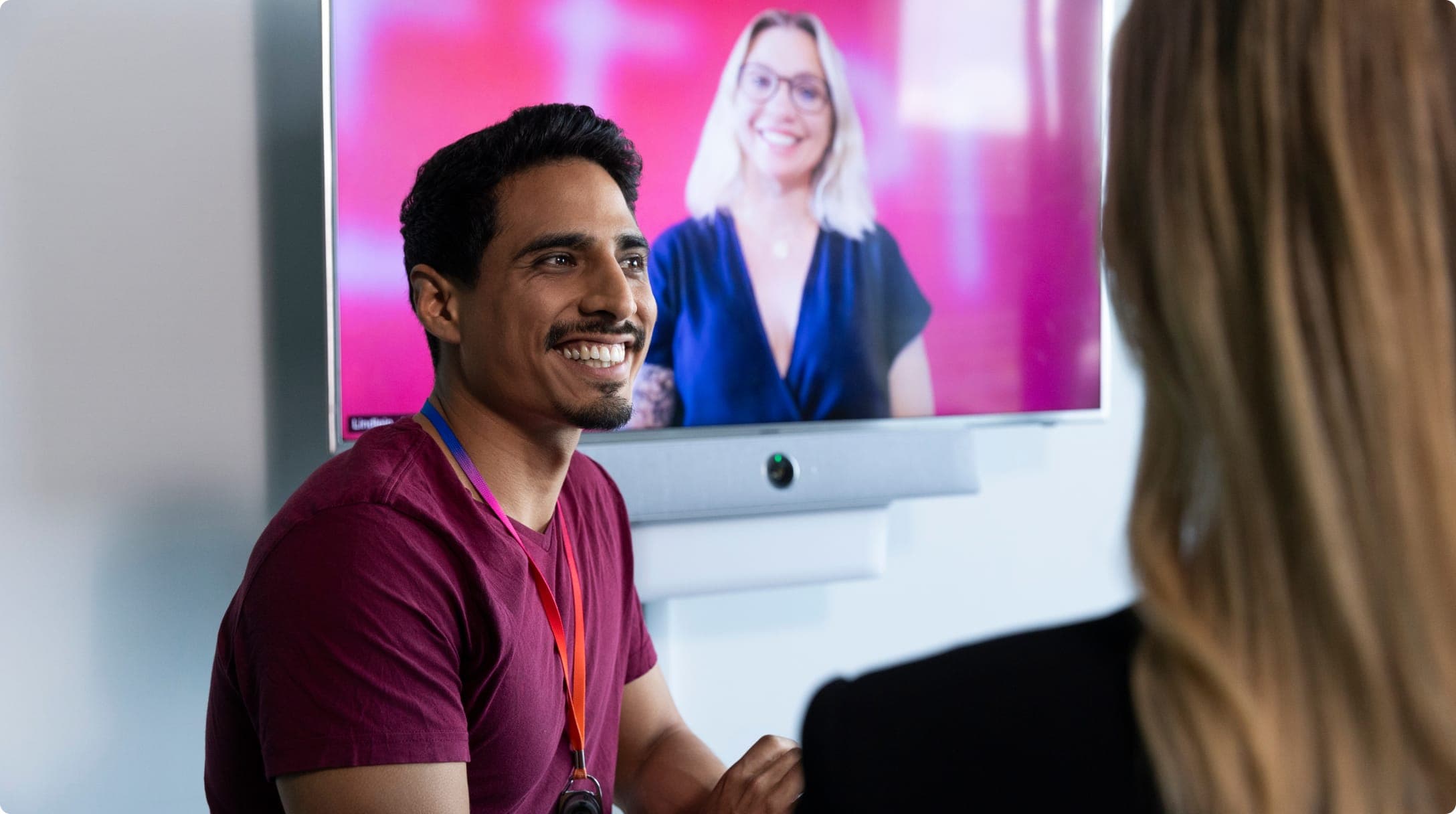 Man smiles whilst on a video call while another colleague listens in a meeting room.