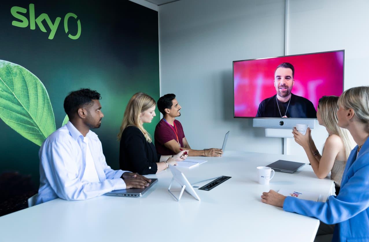 Fünf Personen in einem Besprechungsraum mit „Sky Zero“-Branding im Gespräch mit einem Kollegen, der digital zugeschaltet ist.