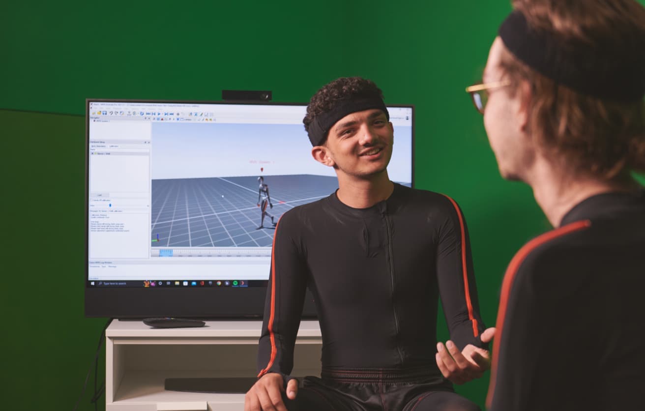 Two men in black motion capture suits talk in front of a screen showing 3D movement tracking software.