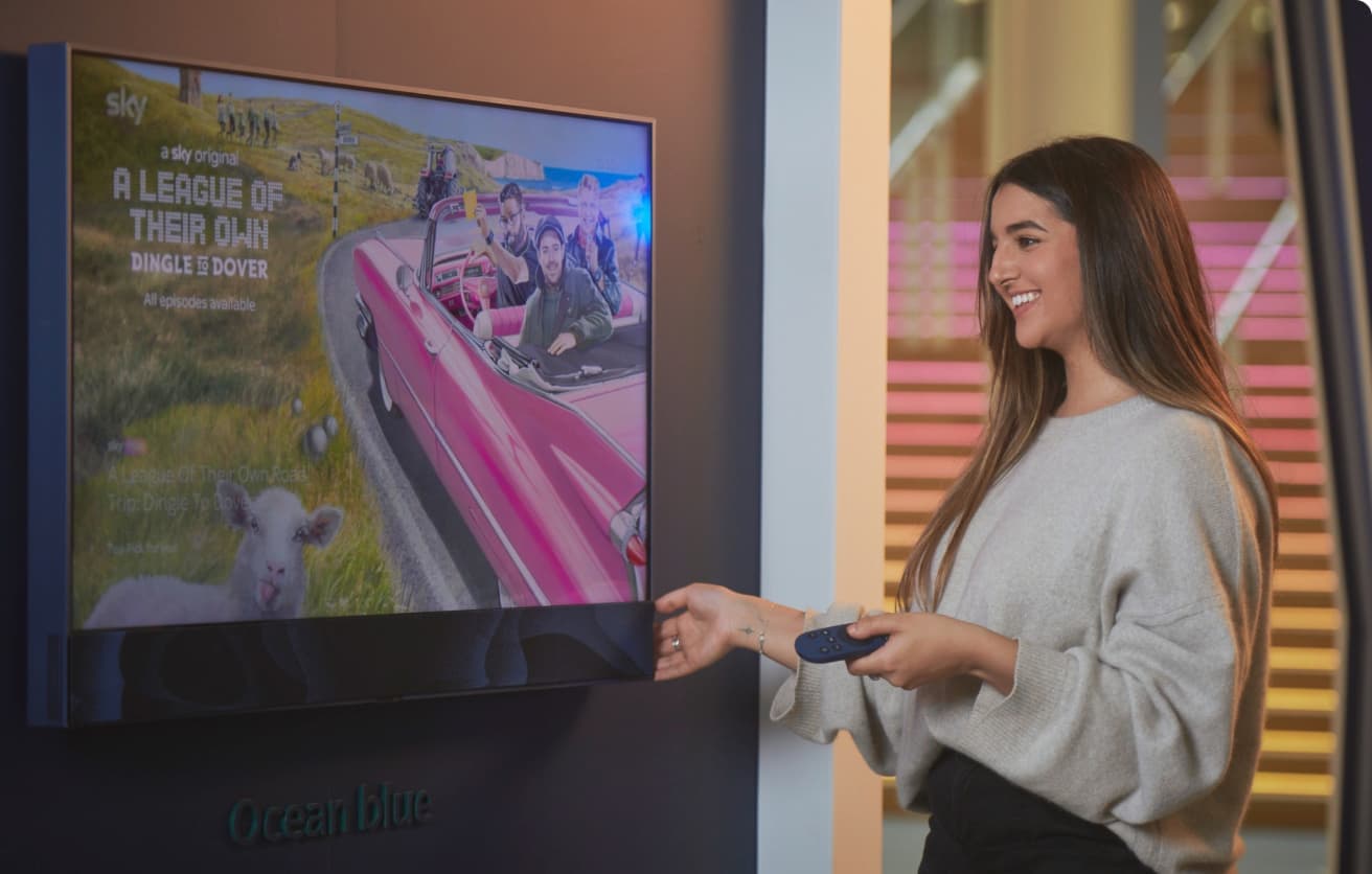 Smiling woman using a remote while standing next to a TV showing a Sky Original episode of A League of Their Own.