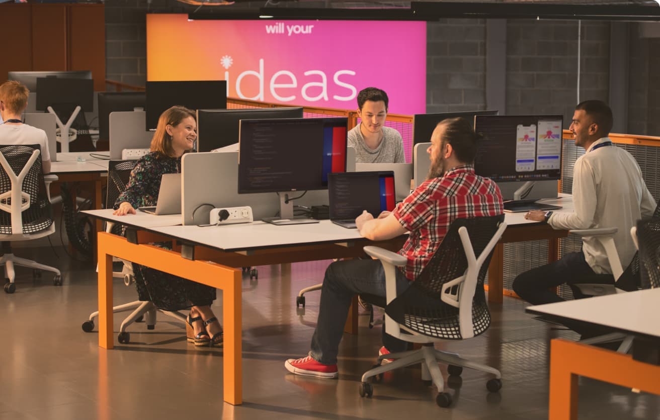 Group of employees collaborating at desks in an open-plan tech office, with a large "ideas" sign in the background.
