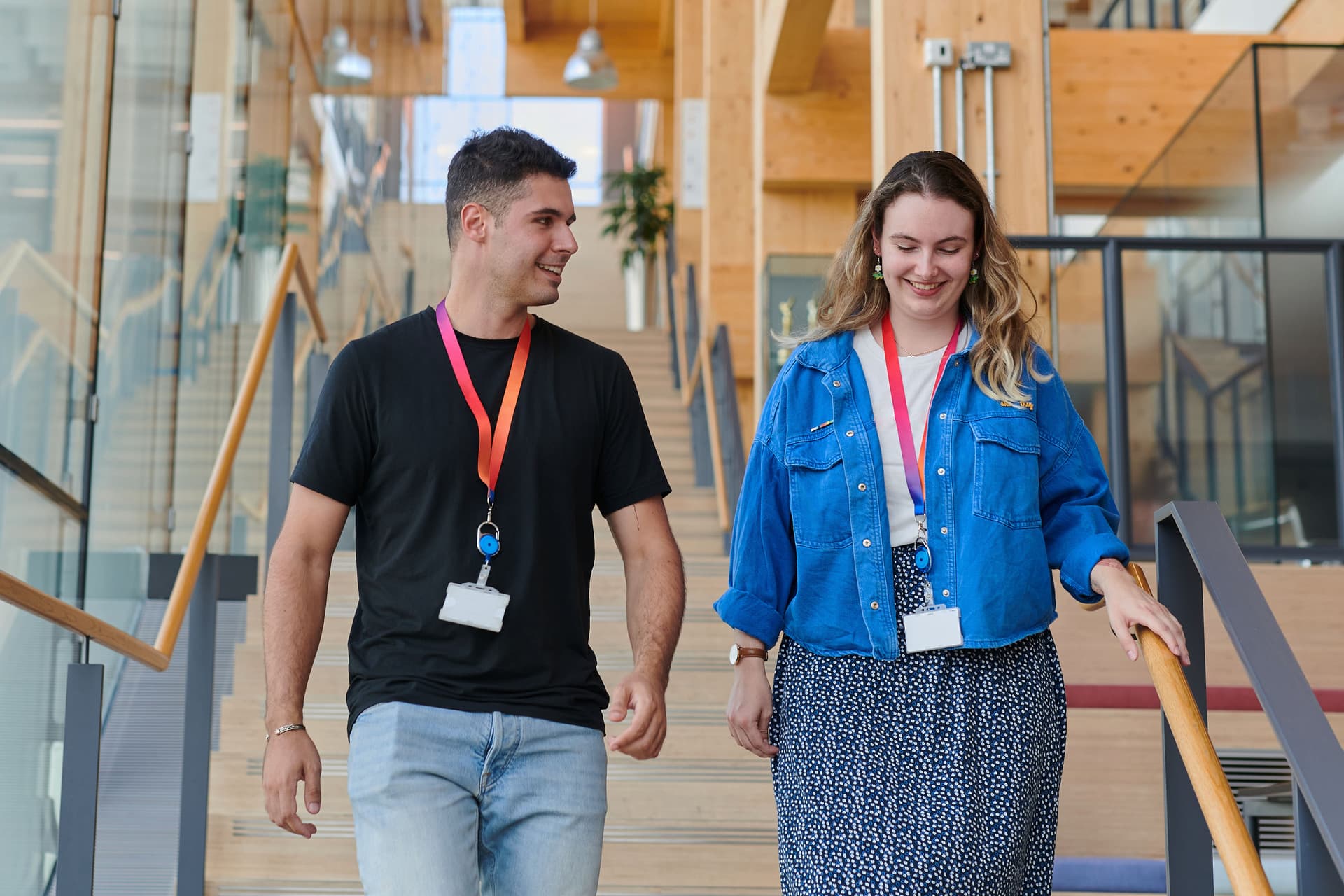 Two coworkers wearing lanyards walk down a wooden staircase in a bright Sky office building.