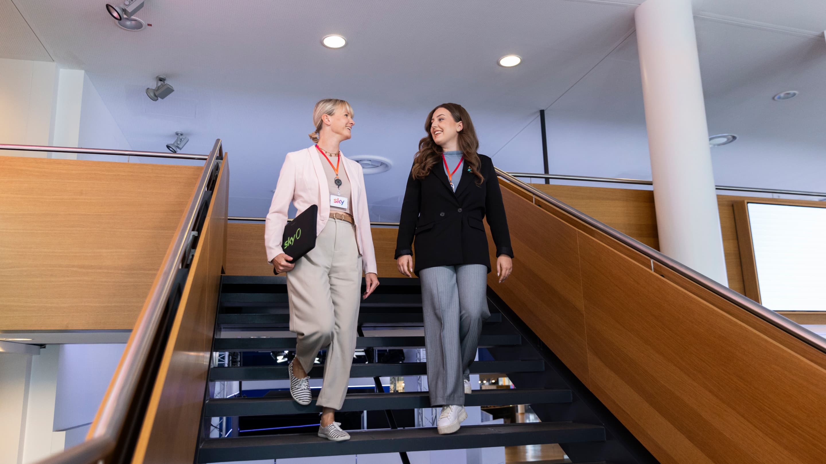 Due donne con badge Sky scendono le scale in un ufficio moderno, sorridenti e con in mano un laptop con il marchio Sky.