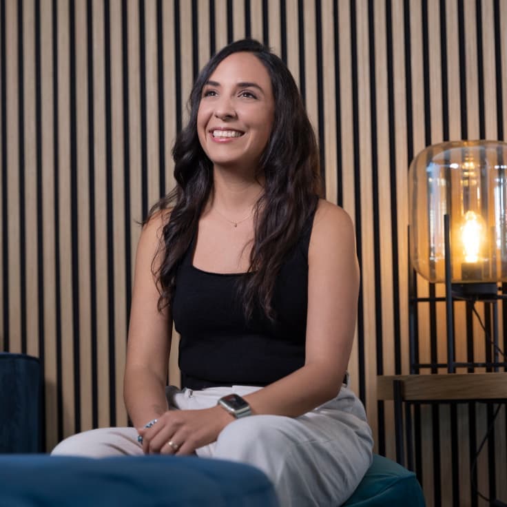 Smiling woman seated on a blue couch, wearing a black top and white pants, in front of a wood-paneled wall and lamp.