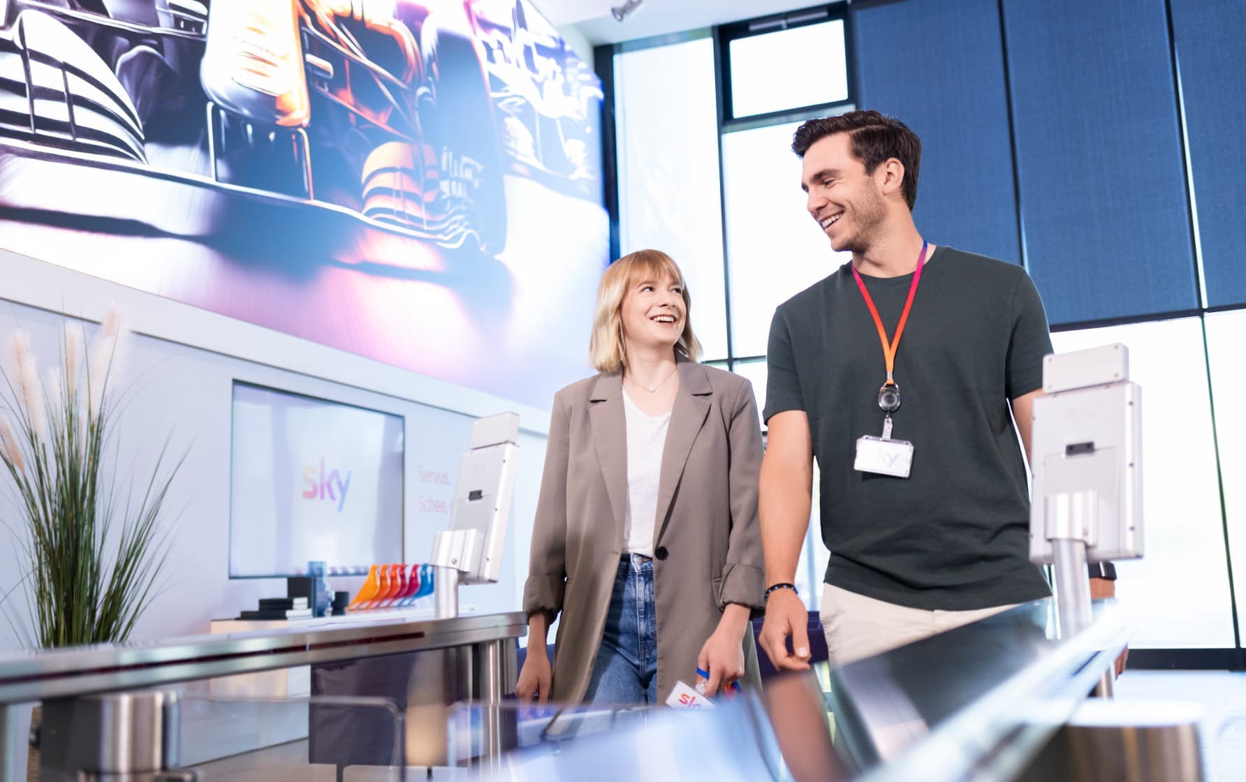 Man and woman wearing Sky badges enter an office lobby with racing artwork on the wall and Sky branding in the background.