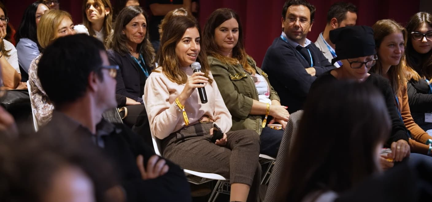 Una donna con un maglione rosa parla al microfono durante un evento aziendale, circondata da colleghi seduti.