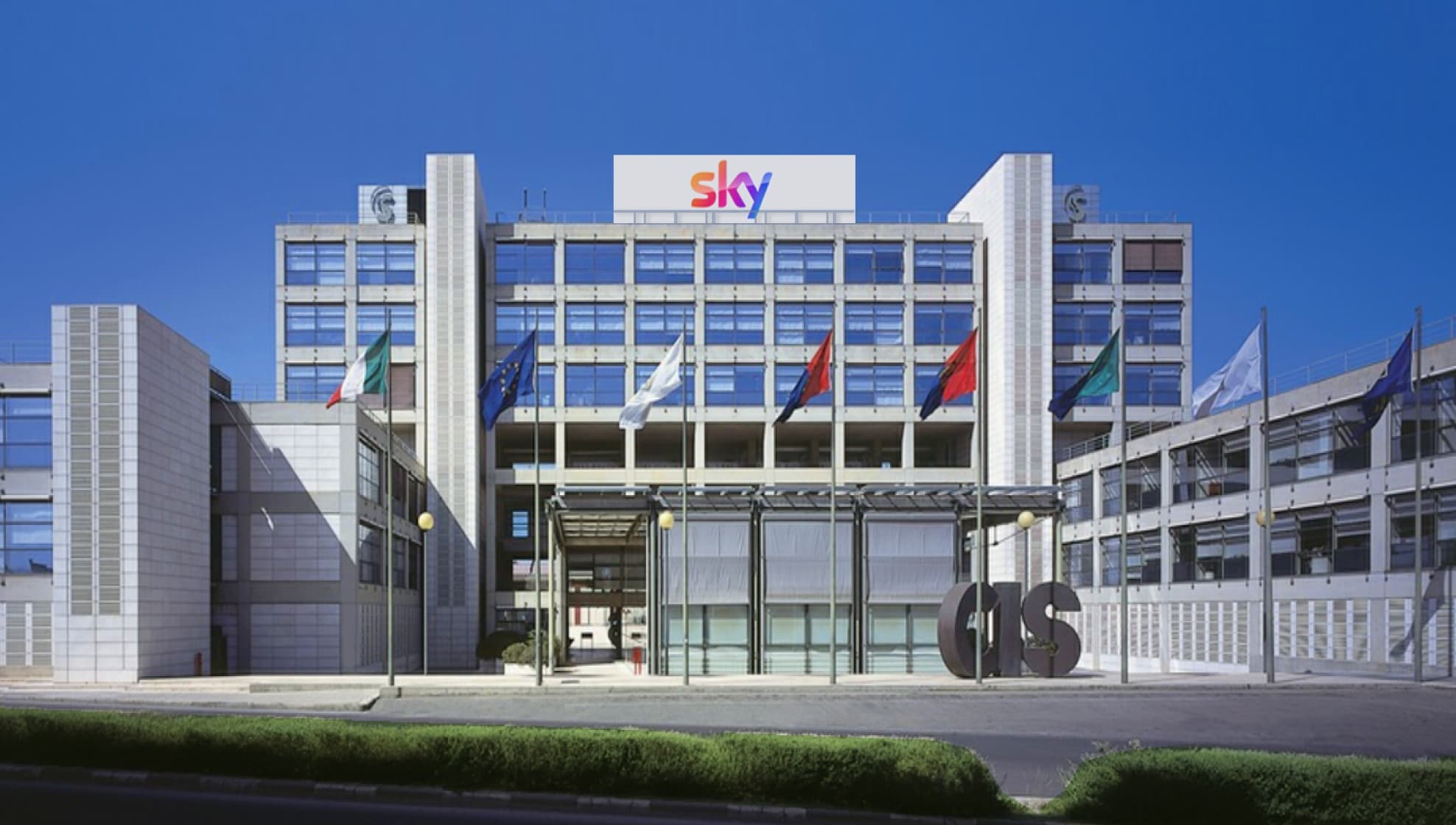 Esterno della sede di Sky Italia a Cagliari con bandiere, sotto un cielo terso e azzurro.