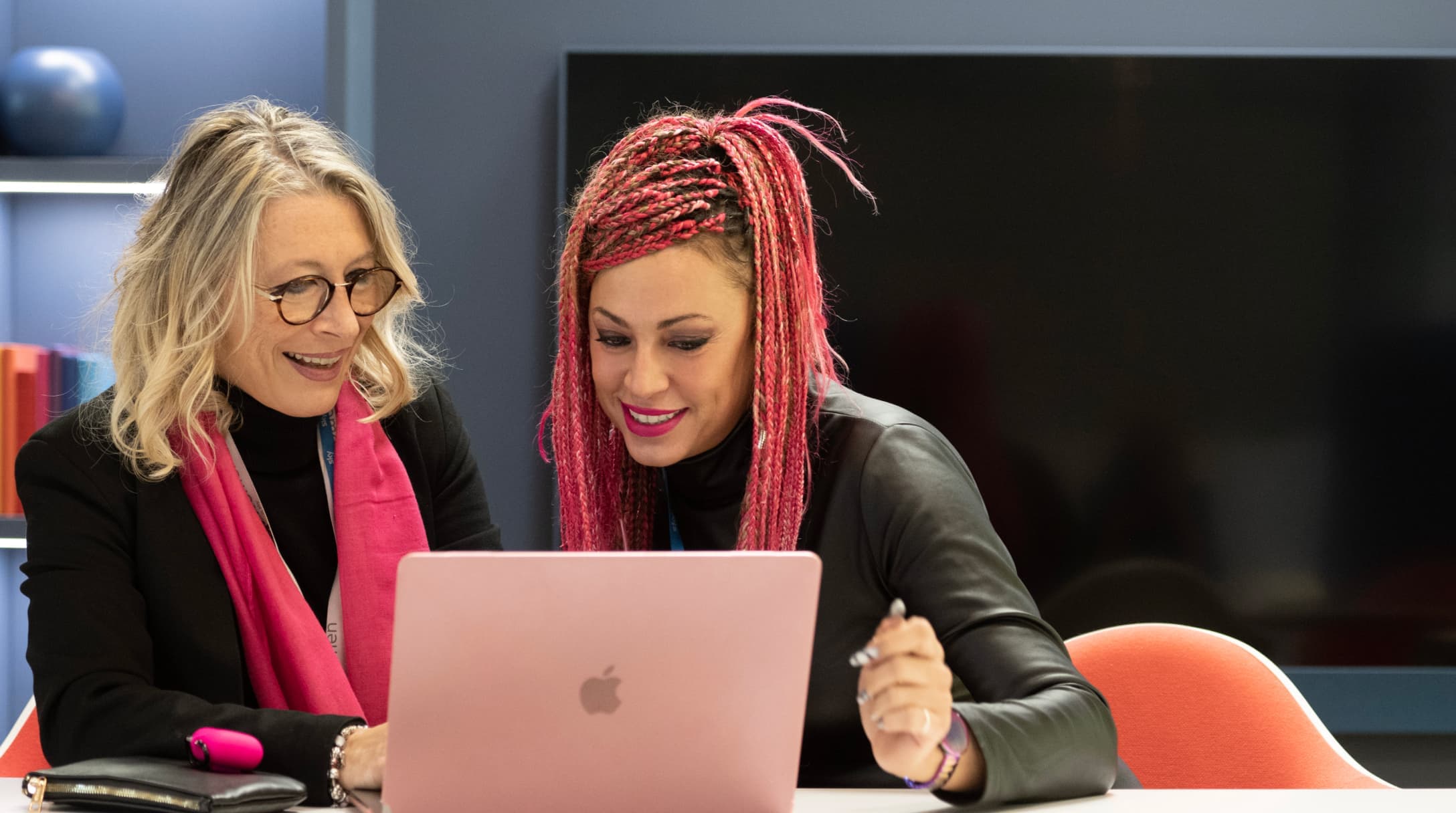 Due donne sorridono mentre guardano un portatile rosa in un moderno ufficio, e sembrano concentrate e collaborative.