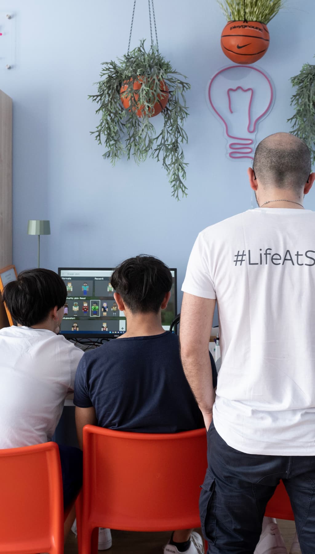 Three people seated at red chairs facing a screen, one wearing a “#LifeAtSky” shirt in a creative, plant-decorated space.