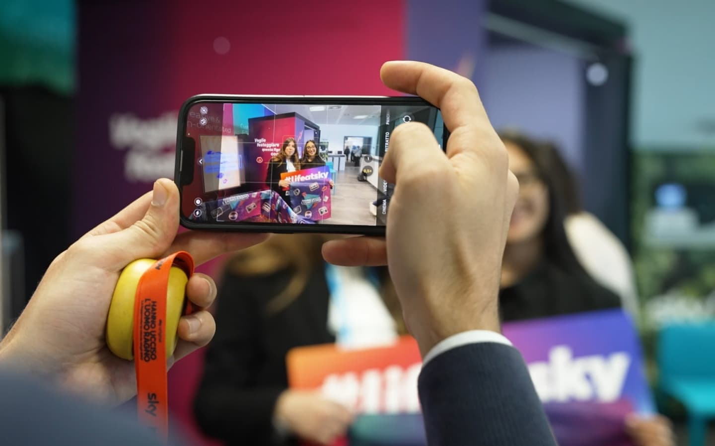 A smartphone captures two smiling Sky employees in a colourful event booth, holding “#lifeatsky” signs.