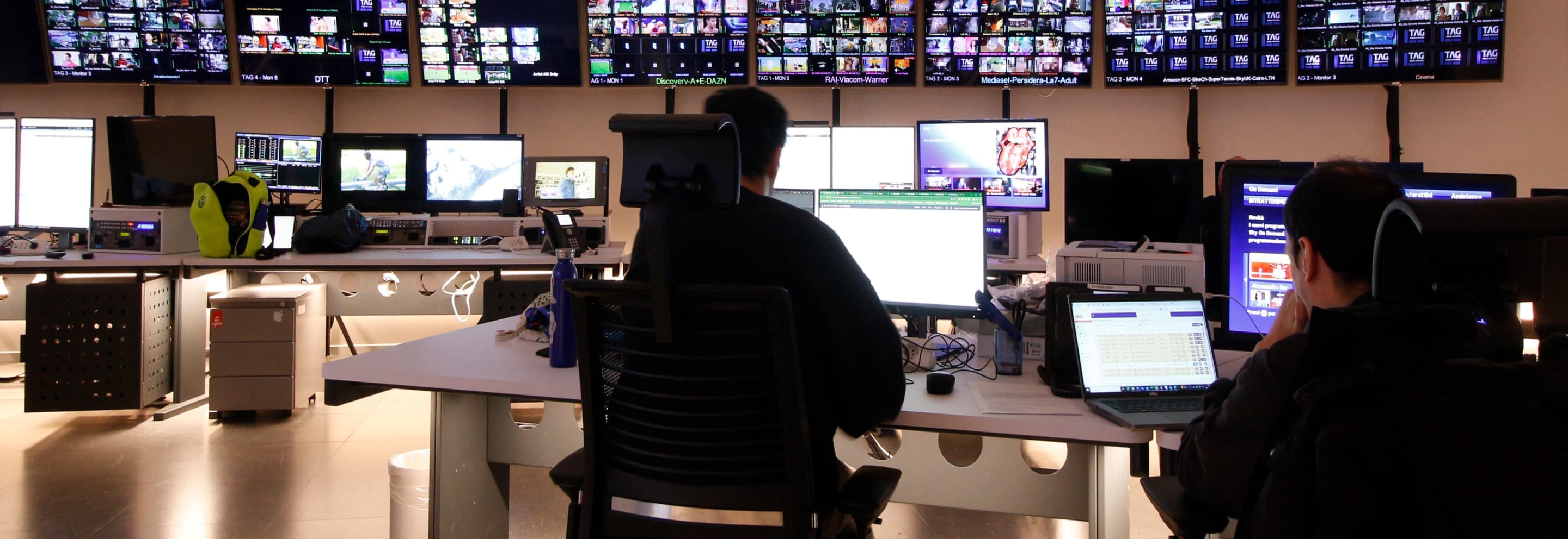 Two people monitor live video feeds and data on multiple screens in a darkened broadcast control room filled with monitors.