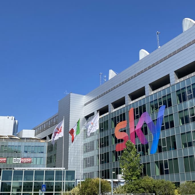 Glass-fronted Sky Italia building in Milan with large Sky logo and Italian and branded flags out front.