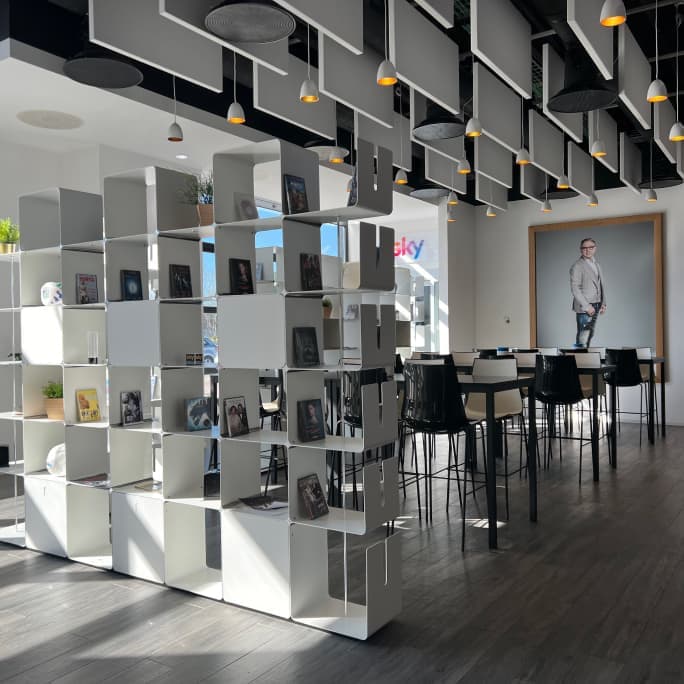 Bright lounge area with bookshelves, tall tables, modern lighting, and a large portrait of a man on the wall.
