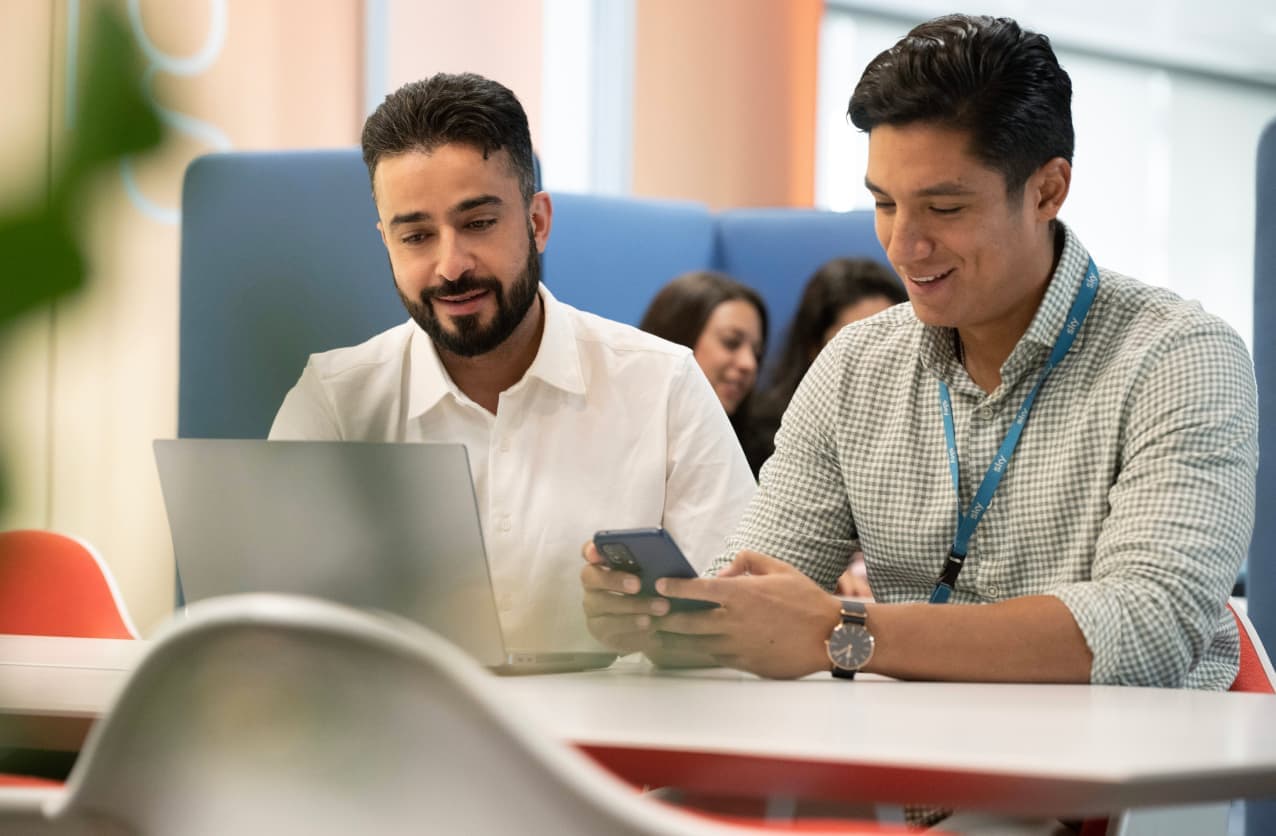 Two men smiling while looking at a phone and laptop in a casual office setting.