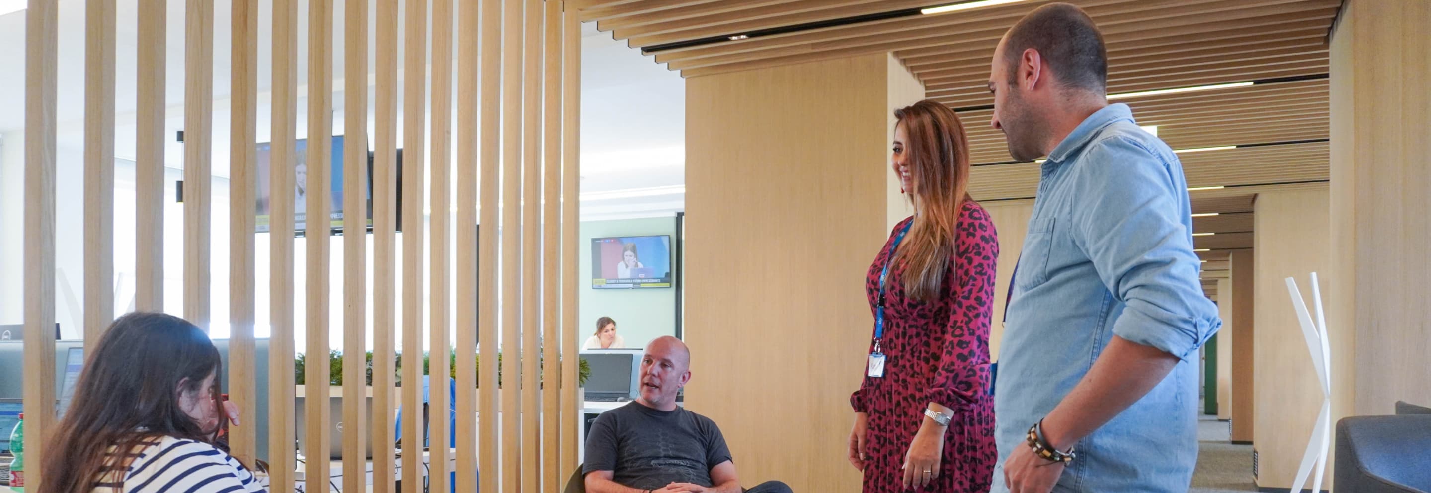 Four colleagues chatting in an open-plan office with wooden paneling, one seated with a laptop.