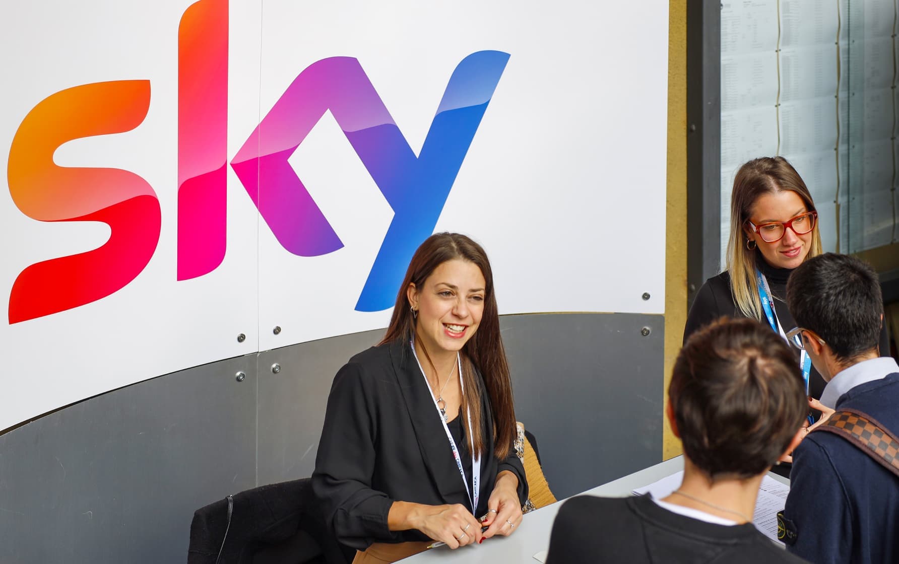 Two women at a Sky-branded desk talking to visitors at a careers or outreach event.