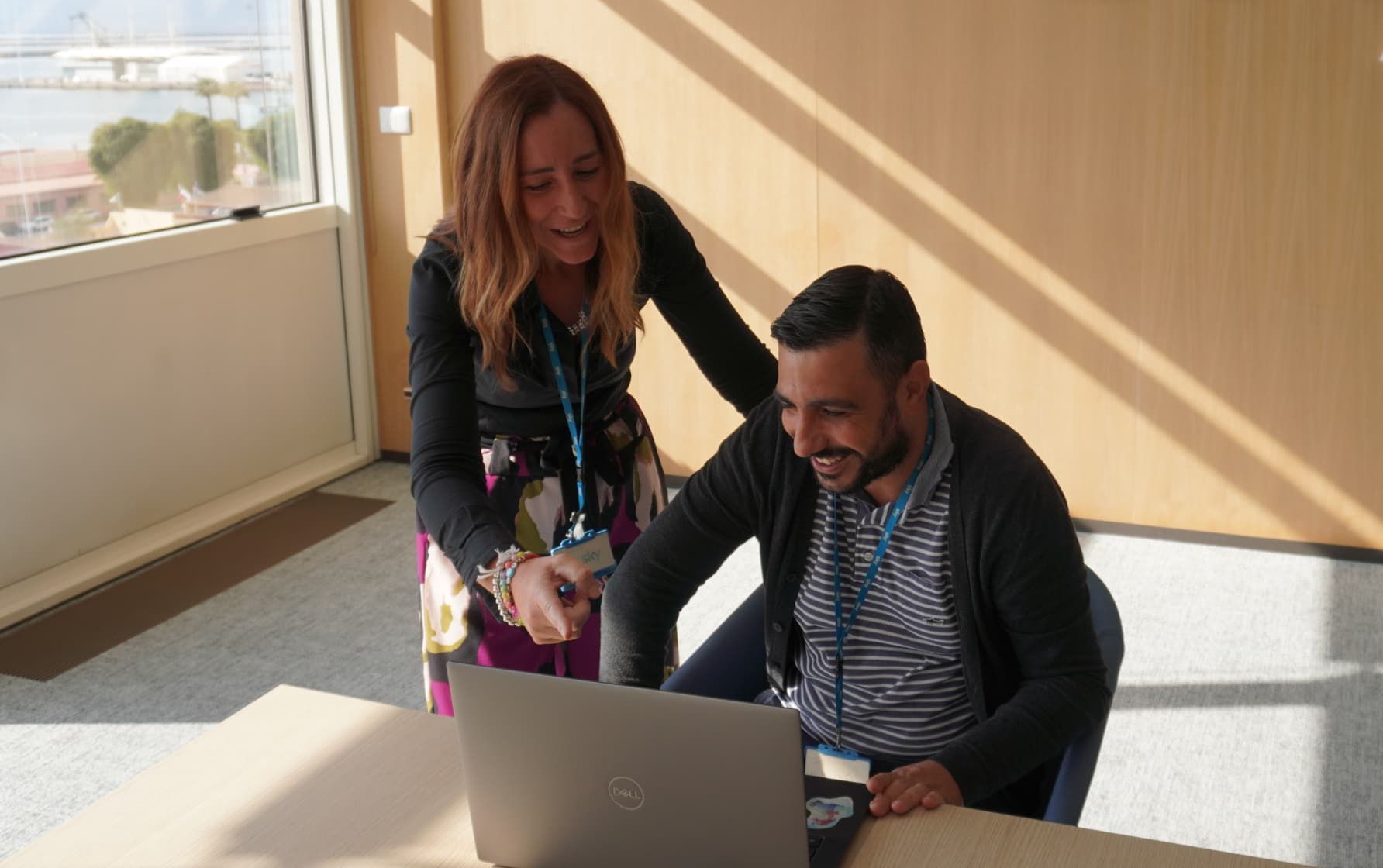 Woman standing and pointing at a laptop while a seated man smiles; both wear Sky lanyards in a sunlit office.