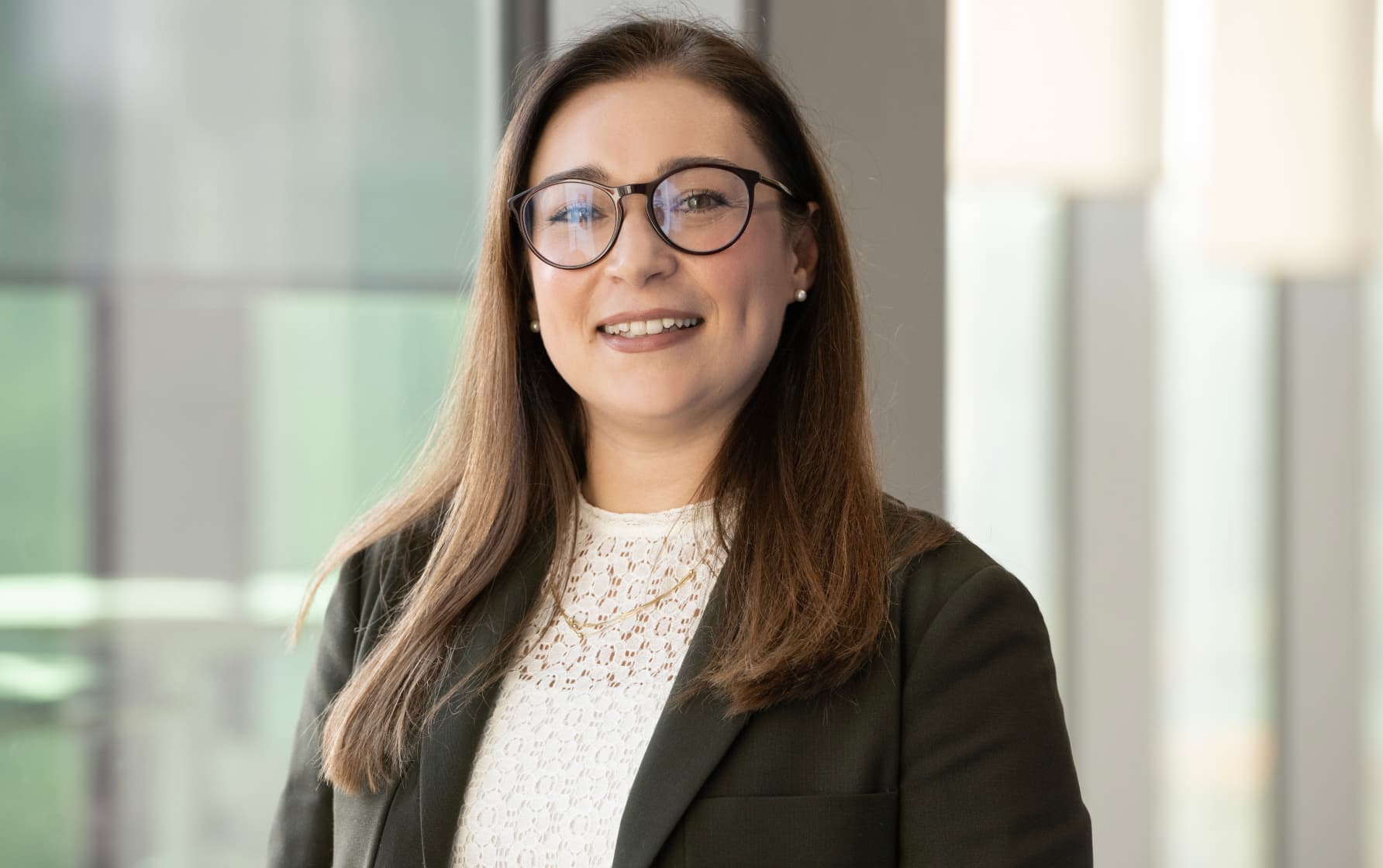 Woman in glasses and blazer smiling in a softly lit office setting with glass-panelled background.