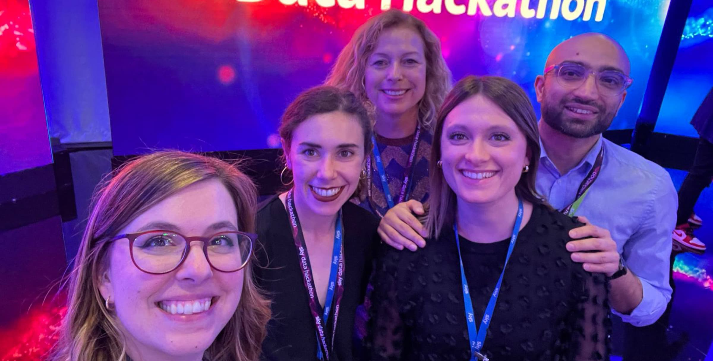 Five smiling colleagues wearing hackathon lanyards take a group selfie in front of a vibrant “Sky Data Hackathon” screen.
