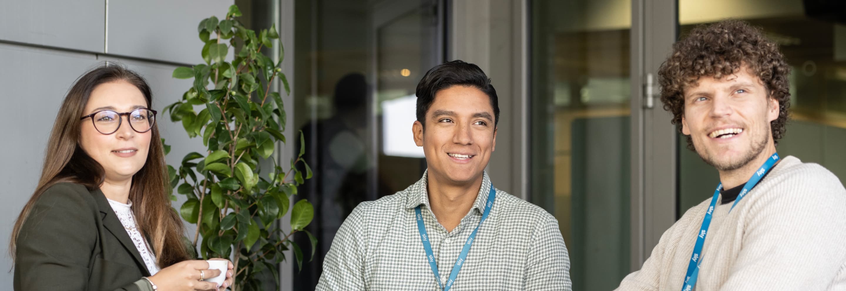 Three Sky employees smiling and chatting near a plant in a modern office setting, all wearing Sky lanyards.