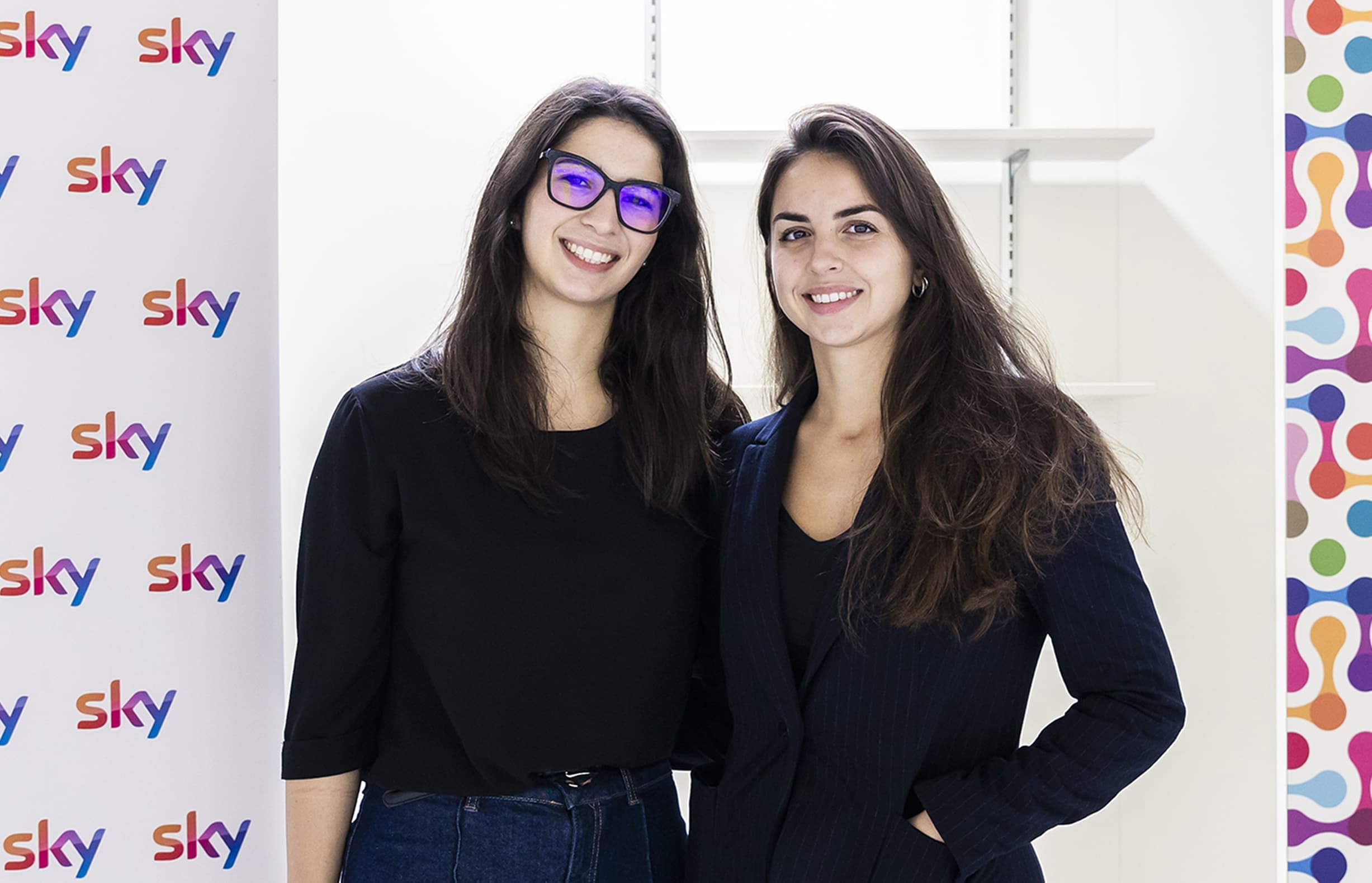 Two women in black outfits stand smiling beside a branded Sky banner in a bright room with a multicolour backdrop.