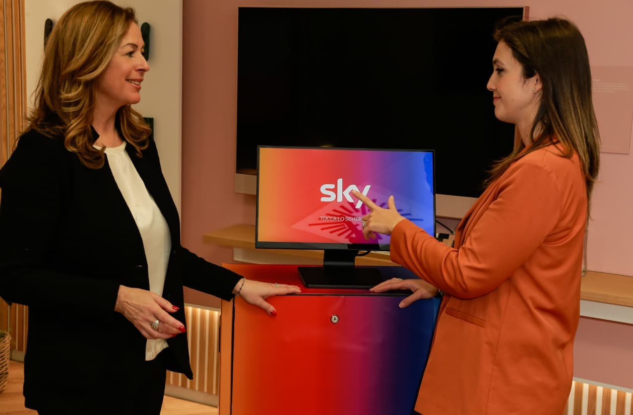 Two women talking at a demo station, one pointing at a small monitor displaying the Sky logo.