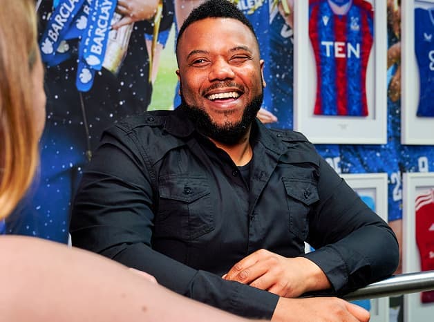 Man in a black shirt smiling while chatting, with framed football shirts in the background.