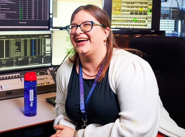 Woman with glasses and Sky lanyard laughing at a control desk surrounded by multiple data monitors.