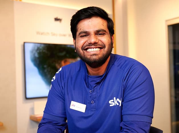 Sky staff member in a blue uniform smiling at the camera inside a Sky retail store.