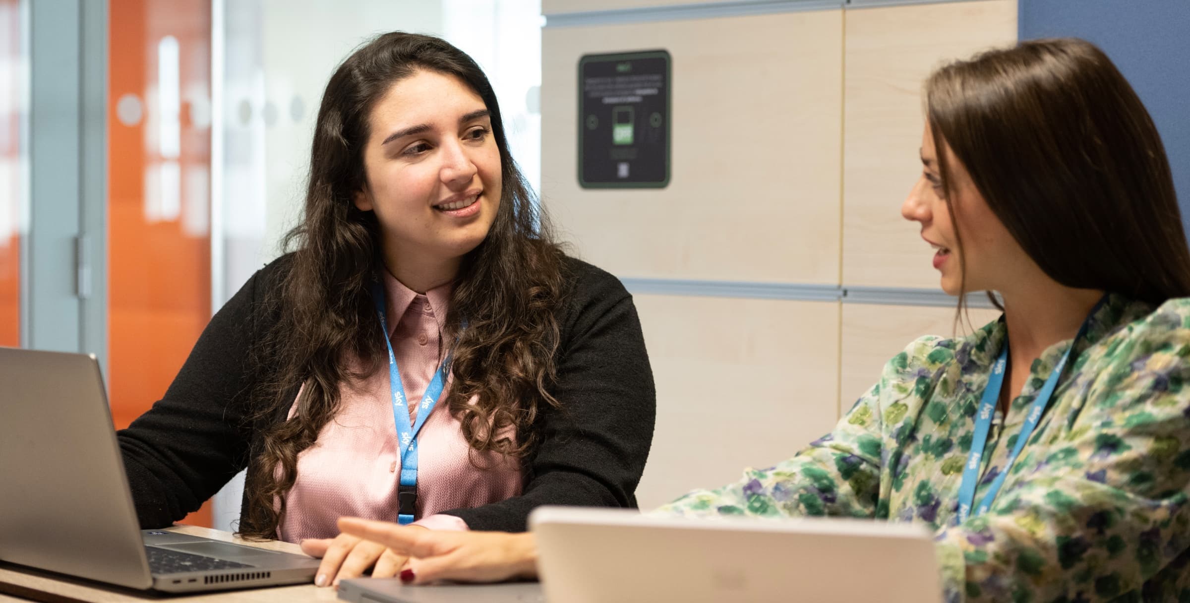 Two women have a friendly discussion while working on laptops in an office with lockers behind them.