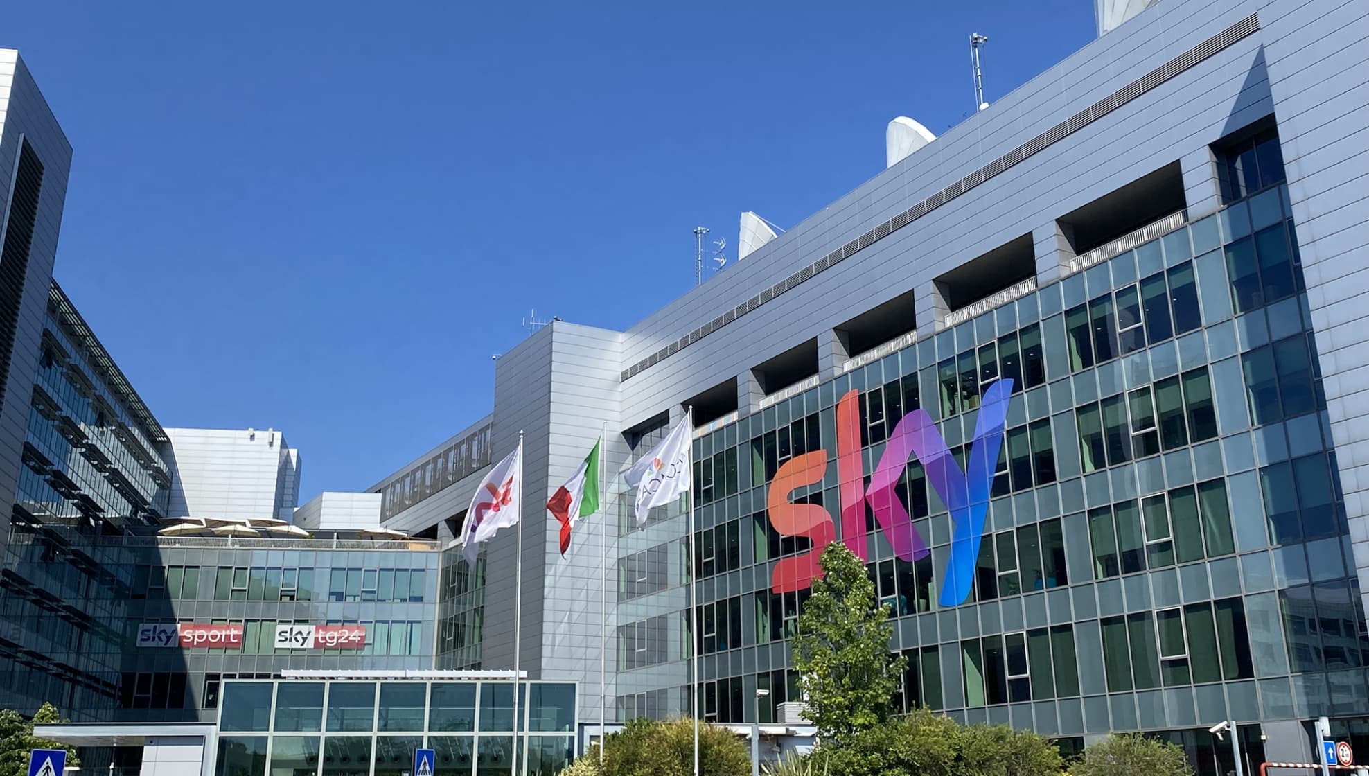 La sede di Sky Italia a Milano con facciata in vetro, grande logo Sky e bandiere italiane davanti alla facciata.